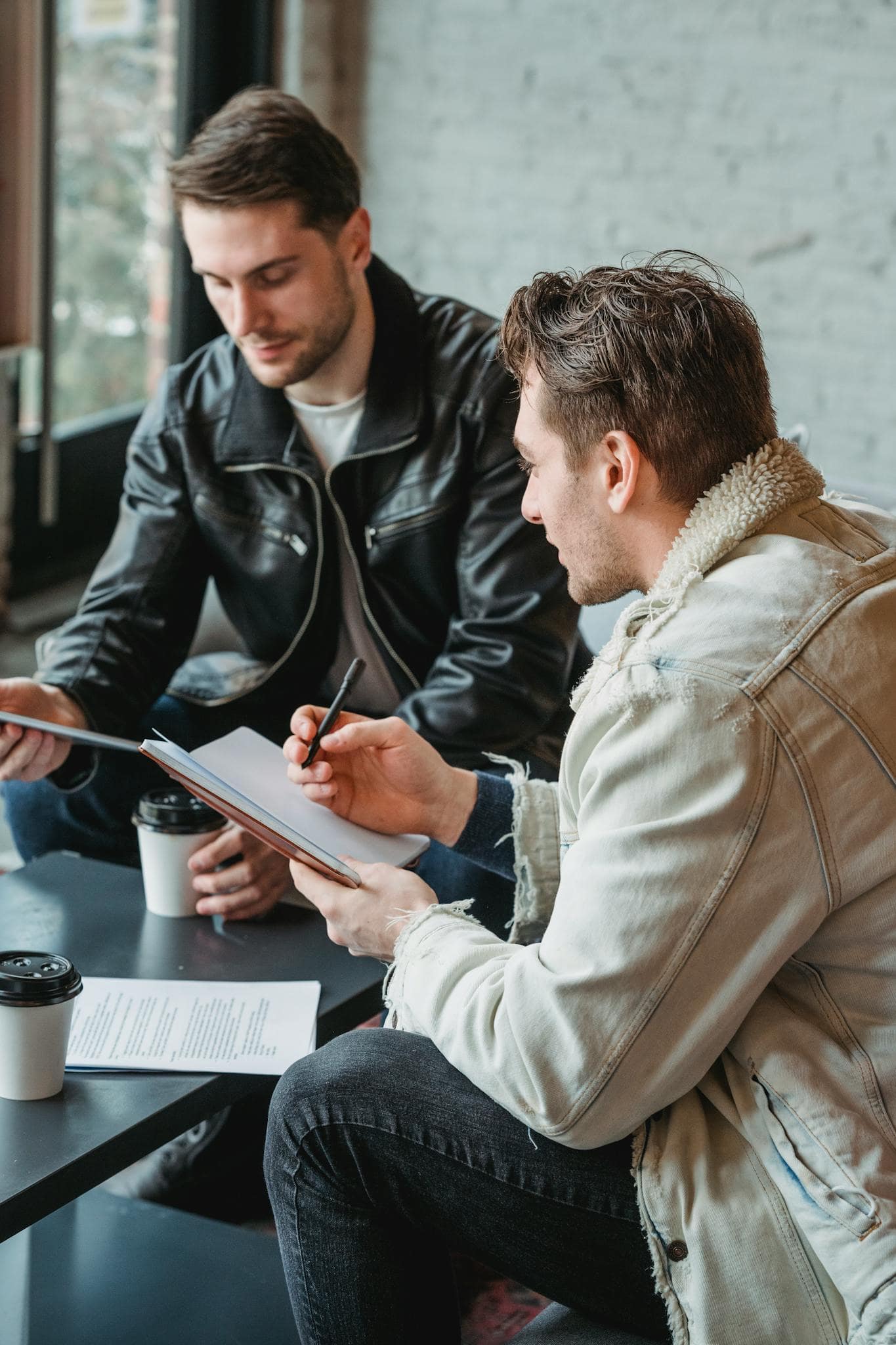 Casual male colleagues sitting together at table and taking notes while drinking coffee in cafe on daytime