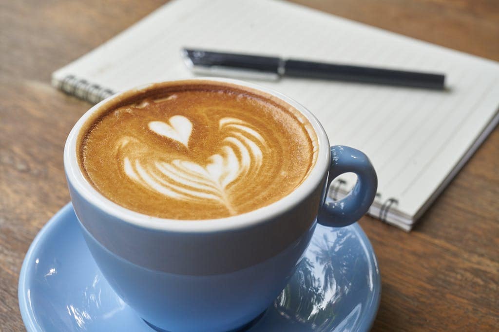 A detailed latte art design in a blue cup next to a notebook and pen on a wooden table.